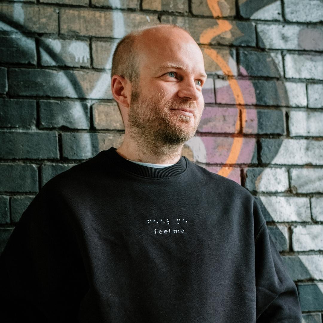 Eine Mann mit kurzen, braunen Haaren steht vor einer Graffitiwand und schaut seitlich neben die Kamera. Er trägt einen schwarzen Pullover mit der weißen Stickung 